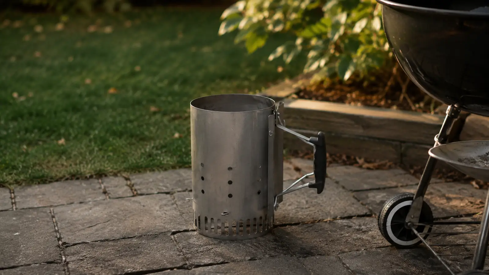 A charcoal chimney starter on a flat stone paver beside a charcoal grill, ready before cooking