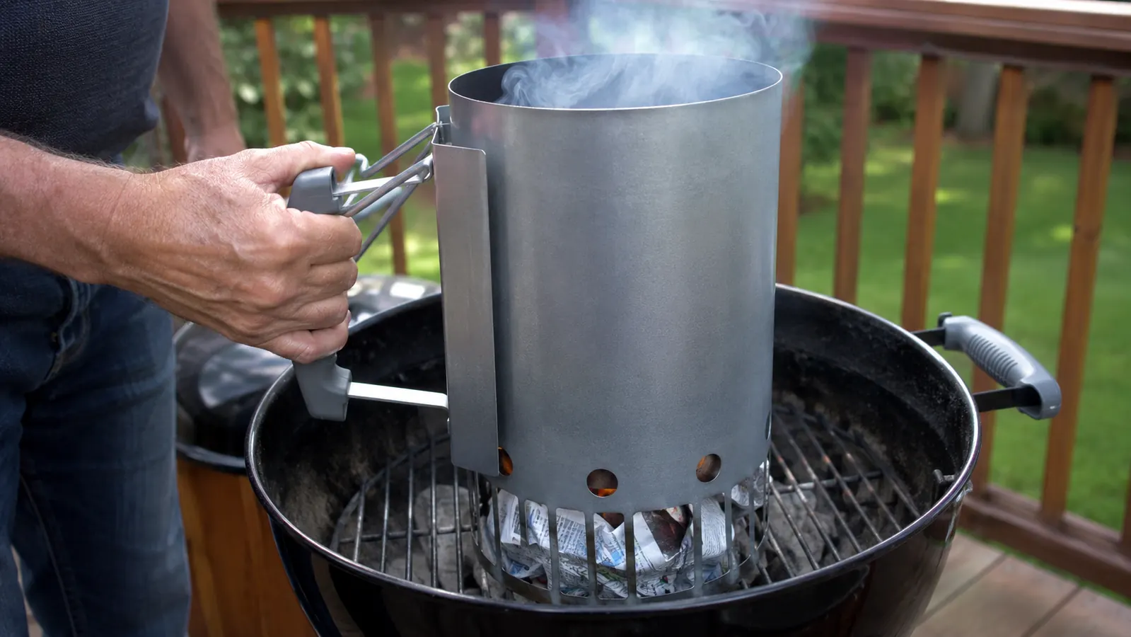 A charcoal chimney starter sitting on a grill grate while a hand holds its handle and thin smoke rises from the bottom.