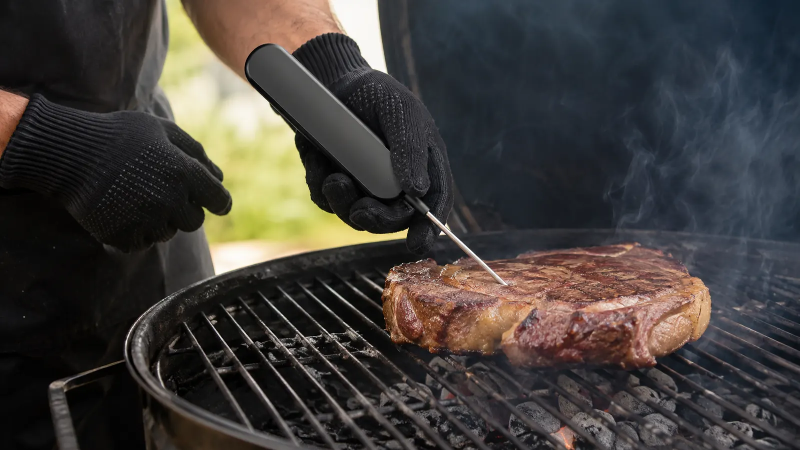 Hand in a heat-resistant glove holding an instant-read meat thermometer over a steak on a grill grate